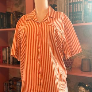 May include: Orange and white striped short-sleeve button-up shirt. The shirt has a collared neckline, a chest pocket, and orange buttons. The vertical stripes are evenly spaced and run the length of the shirt. The shirt is displayed on a mannequin.