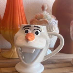 May include: A white ceramic mug shaped like a character face with a prominent orange carrot nose, black eyebrows, and a wide open mouth. The mug features a smooth, glossy finish, a sturdy handle on the right side, and a circular base for stability.