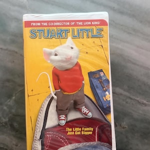 May include: A Stuart Little VHS tape featuring a white mouse wearing a red sweater and brown pants standing on a red and white sneaker. The cover includes the text 'FROM THE CO-DIRECTOR OF THE LION KING', 'STUART LITTLE', and 'The Little Family Just Got Bigger'.