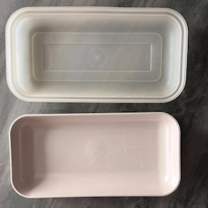 May include: Two rectangular plastic containers. The top container is translucent with a lid, and the bottom container is a solid light pink. Both containers are empty and have rounded corners.