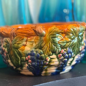 May include: A ceramic bowl with a detailed grape and leaf design. The bowl features a vibrant color palette of orange, green, blue, and purple. The rim has a textured, raised design, and the bowl sits on a dark surface.