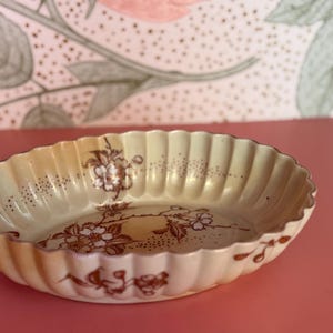 May include: An oval, cream-colored ceramic dish with a fluted rim. The dish features a floral design in shades of brown and is set against a pink surface. The background has a floral pattern.