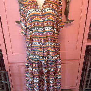 May include: A two-piece set consisting of a short-sleeved button-down shirt and a matching long skirt. The fabric features a repeating geometric pattern in shades of orange, purple, teal, and white, with triangular and curved abstract shapes.