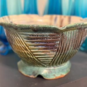 May include: A handcrafted ceramic bowl with a textured exterior. The bowl features a green, brown, and tan color palette. The exterior has a ribbed design. The bowl sits on a small, round base.