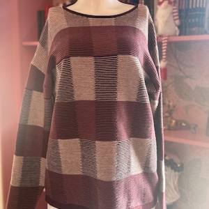 May include: A burgundy, tan, and brown checkered sweater with a crew neck and long sleeves. The sweater is displayed on a mannequin. The background includes a pink bookshelf and decorative items.