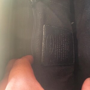 May include: Inside a black handbag, showing a zipped section and a leather label. The label has embossed text and a logo. The bag's lining is dark, and a hand is partly visible.