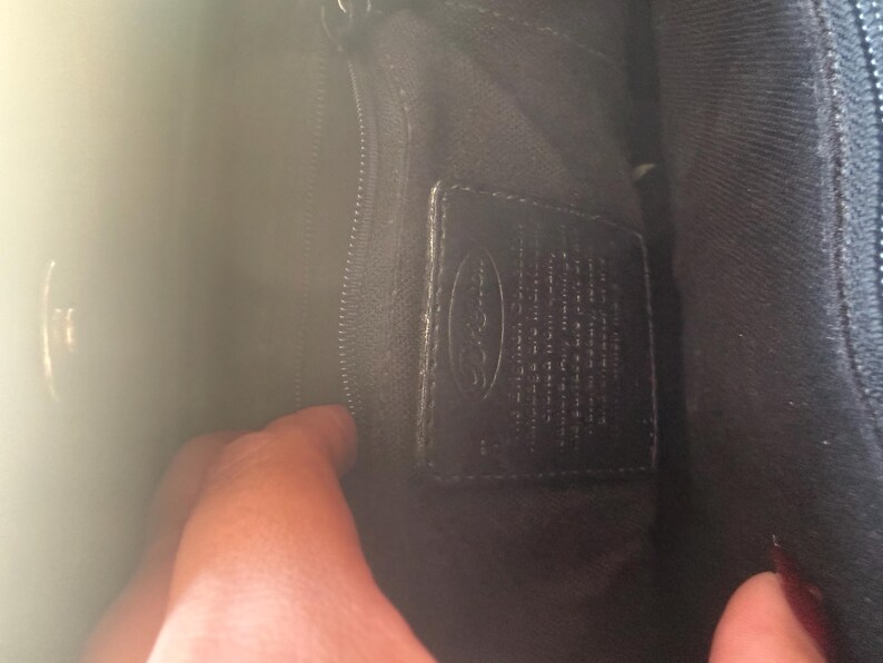 May include: Inside a black handbag, showing a zipped section and a leather label. The label has embossed text and a logo. The bag's lining is dark, and a hand is partly visible.