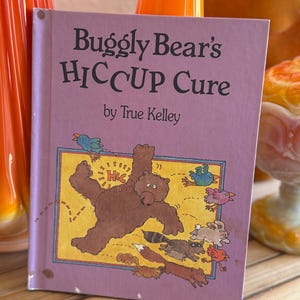 May include: A purple book titled "Buggly Bear's HICCUP Cure" by True Kelley. The cover features a cartoon illustration of a brown bear with a speech bubble saying "Hic" and various animals surrounding it.