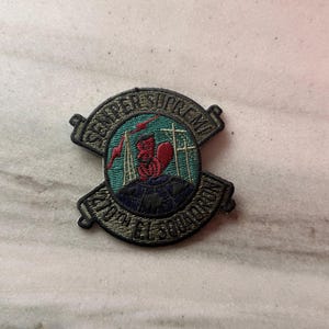 May include: A circular embroidered patch featuring a red squirrel sitting on a dark blue globe. The background is teal with a red lightning bolt and two radio towers. The black border text reads SEMPER SUPREMA and 210TH EL SQUADRON.