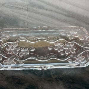 May include: A rectangular clear glass serving tray featuring a wavy center design and frosted floral motifs. The tray has decorative scrollwork at both ends, with a smooth, transparent finish and raised, etched flower patterns across the surface.