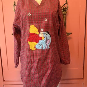 May include: A long-sleeve V-neck tunic featuring a red and black plaid pattern. The front displays an embroidered applique of Winnie the Pooh and Eeyore, accented with small white embroidered snowflakes scattered above the characters.