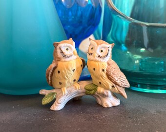 May include: A ceramic figurine featuring two owls perched on a branch. The owls have brown and yellow plumage with black spots and detailed facial features. The branch has green leaves, and the background includes blue glass vases.
