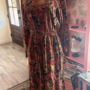 May include: A long-sleeved dress with a floral pattern in shades of red, brown, and black. The dress has a collared neckline, an elastic waist, and a flowing skirt. The fabric appears to be a soft, lightweight material.
