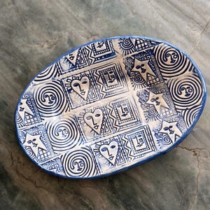 May include: An oval ceramic dish with a blue rim and a white interior. The interior is decorated with blue geometric patterns, faces, hearts, and sun-like designs. The dish is likely handmade and suitable for serving food or as a decorative piece.