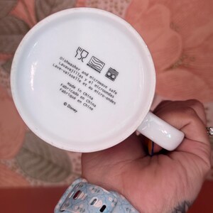 May include: White ceramic mug with a handle, featuring text indicating it's dishwasher and microwave safe. The text includes "Made in China" and the Disney copyright symbol. The mug is held by a person.