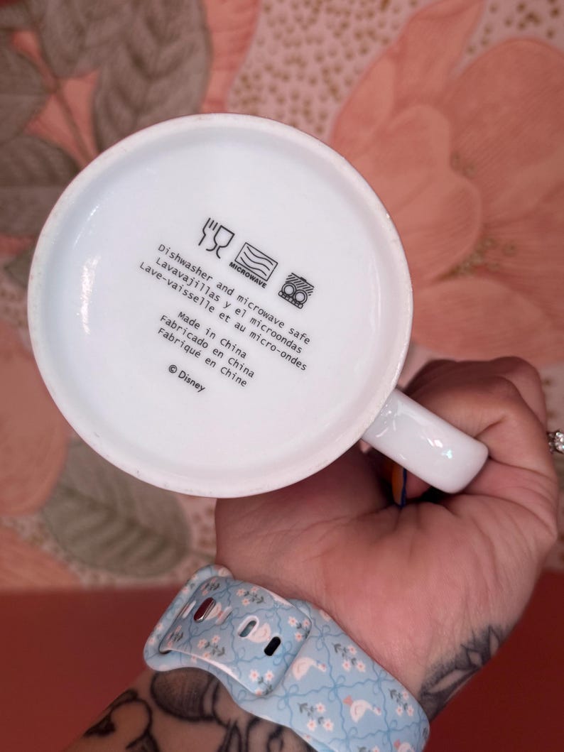 May include: White ceramic mug with a handle, featuring text indicating it's dishwasher and microwave safe. The text includes "Made in China" and the Disney copyright symbol. The mug is held by a person.