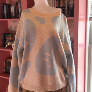 May include: A light peach-colored sweater with a blue and peach pattern. The sweater is displayed on a mannequin. The sweater has a round neckline and long sleeves. The pattern is a repeating design of abstract shapes.
