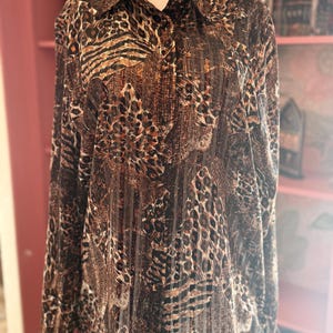 May include: A button-down shirt with a brown and black animal print design. The long-sleeved shirt features a collared neckline and a mix of leopard and zebra patterns. The fabric has a slightly sheer appearance.