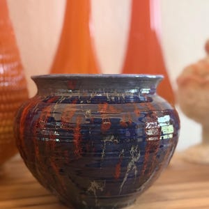 May include: A small, round ceramic pot with a wide, flared rim. The exterior features a glossy, deep blue glaze with vertical streaks of burnt orange and metallic silver accents, creating a textured, layered appearance across the surface.