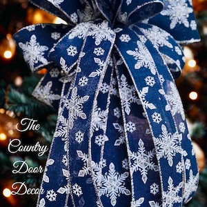 Op de afbeelding: Een grote, decoratieve strik gemaakt van marineblauw lint, versierd met witte sneeuwvlokpatronen en zilveren accenten. De strik hangt aan een kerstboom, met de tekst "The Country Door Decor" zichtbaar.