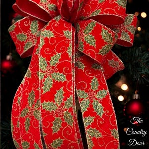Pode incluir: Um grande laço de Natal vermelho festivo com um padrão de folhas de azevinho em verde e dourado. O laço é feito de uma fita larga e é exibido contra um fundo desfocado de uma árvore de Natal e luzes. O texto The Country Door Decor é visível.