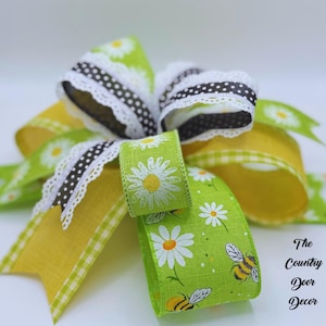 Op de afbeelding: Decoratieve strik met gele, groene en zwarte linten. Het groene lint heeft een patroon van madeliefjes en bijen. Het zwarte lint heeft witte stippen en een witte kanten rand. Geschikt voor knutselprojecten.