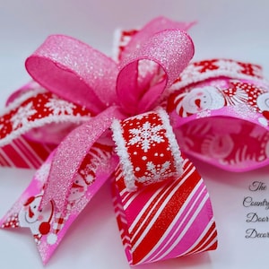 Pode incluir: Um laço de Natal festivo com fitas rosa e vermelhas. O laço inclui uma fita rosa brilhante, uma fita vermelha com flocos de neve brancos e uma fita com um design de Papai Noel. Um item decorativo para as festas.