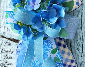 May include: A blue hydrangea floral arrangement with various shades of blue ribbons and accents. The arrangement includes a gingham ribbon, a floral print ribbon, and a glitter ribbon. The text "The Country Door Decor" is visible.