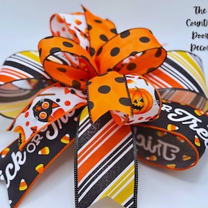 Pode incluir: Um laço decorativo de Halloween com bolinhas laranja, listras e padrões temáticos. O laço inclui fitas com abóboras, doces de milho e as frases "Trick or Treat". The Country Door Decor está impresso na lateral.