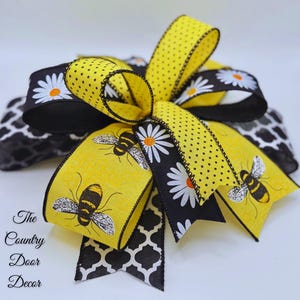 May include: A decorative bow featuring yellow and black patterned ribbons. The bow has a yellow ribbon with black polka dots and bee illustrations. Other ribbons have daisy and geometric patterns. The text "The Country Door Decor" is visible.