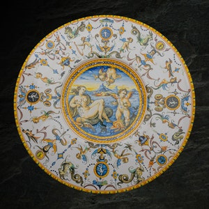 May include: A white ceramic plate with a blue and yellow border. The center of the plate features a colorful painting of a woman and a cherub in a seascape. The plate is decorated with a variety of floral and geometric designs.
