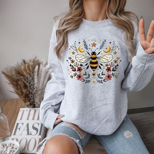May include: A grey sweatshirt featuring a graphic of a bee surrounded by flowers and stars. The bee is yellow and black with brown stripes. The flowers are pink, red and yellow.
