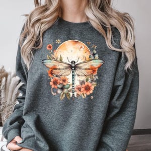 May include: Dark gray sweatshirt featuring a watercolor design of a dragonfly with orange and blue wings, surrounded by flowers and a sun. The design is centered on the front of the sweatshirt.