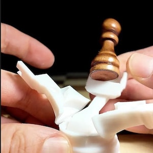 May include: A white plastic chess piece with a brown wooden pawn on top. The pawn is being held by a hand and is about to be placed on the chess piece.
