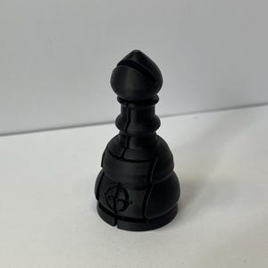 Può includere: Un pezzo degli scacchi nero con una cima arrotondata e un disegno circolare sulla base.