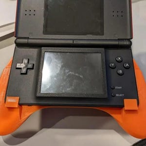 May include: A black Nintendo DS handheld gaming console with an orange 3D printed controller attachment. The console has a black screen and buttons labeled X, Y, A, B, Start, and Select.