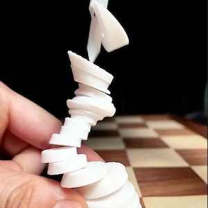 Op de afbeelding: Een wit schaakstuk wordt in elkaar gezet op een houten schaakbord. Het stuk bestaat uit meerdere ronde stukken die in elkaar passen om een torenvormige structuur te vormen.