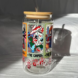 May include: A clear glass tumbler with a bamboo lid and a straw. The tumbler features a graphic of four horror movie characters with blood splatters and the text "The Camper" and "The K".