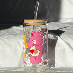May include: A clear glass tumbler in a can shape, with a bamboo lid and glass straw. The tumbler is decorated with Care Bear characters and floral patterns. The design includes a pink Care Bear with a heart. A charming and playful drinking vessel.