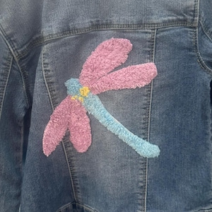 May include: A denim jacket with a large, embroidered dragonfly design on the back. The dragonfly features pink wings, a blue body, and yellow accents, creating a colorful and textured detail on the jacket.