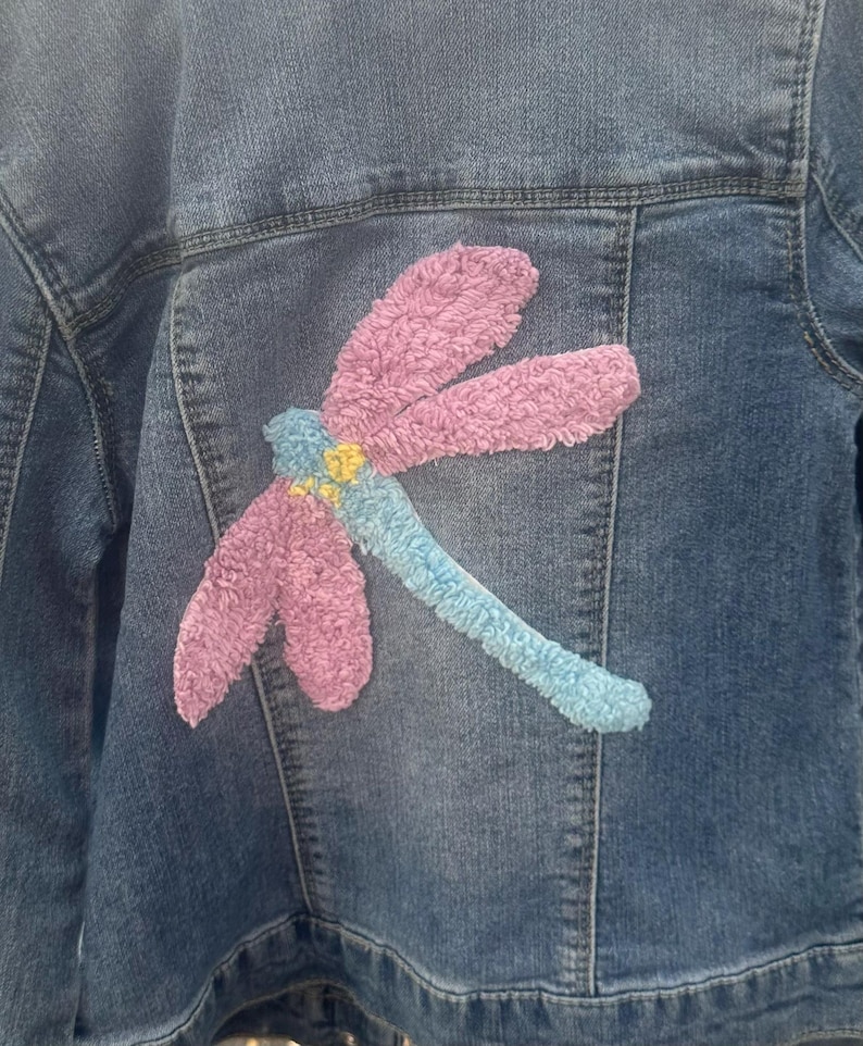 May include: A denim jacket with a large, embroidered dragonfly design on the back. The dragonfly features pink wings, a blue body, and yellow accents, creating a colorful and textured detail on the jacket.