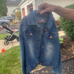 May include: A blue denim jacket with copper-colored buttons and a collar. The jacket is hanging on a wooden hanger. The jacket has two chest pockets and a label on the back. The jacket is a medium wash denim.