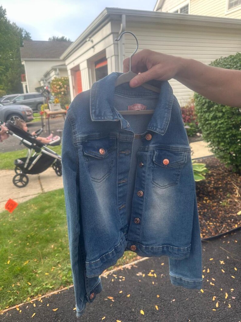 May include: A blue denim jacket with copper-colored buttons and a collar. The jacket is hanging on a wooden hanger. The jacket has two chest pockets and a label on the back. The jacket is a medium wash denim.