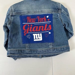 May include: A blue denim jacket with a patch featuring the words "New York Giants" in red and white, with the team's logo. The jacket is hanging on a metal hanger.