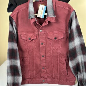 Pode incluir: Jaqueta jeans bordô com mangas xadrez cinza e preto. A jaqueta tem dois bolsos no peito, fechos de botão e gola xadrez cinza. Uma camiseta preta está pendurada atrás da jaqueta.