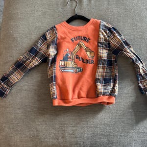 Pode incluir: Camiseta laranja de manga comprida com mangas xadrez. A frente apresenta um gráfico de um veículo de construção e as palavras "FUTURE BUILDER". A camiseta está pendurada em um cabide preto.
