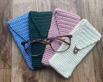 May include: A collection of handmade crochet eyeglass cases in blue, green, pink, and cream. A pair of eyeglasses rests on top of the cases. The cases have a flap closure with a wooden button.