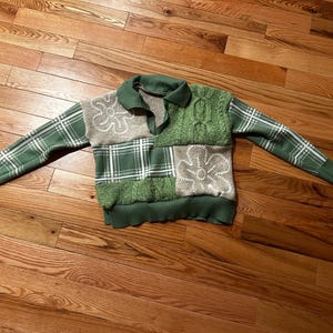 May include: A patchwork sweater with a green and white plaid pattern, cable knit, and floral embroidery. The sweater has a green collar and cuffs. The design incorporates various textures and patterns, creating a unique and stylish garment.