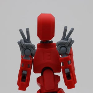 May include: A red and gray robot toy with articulated arms and legs. The robot is making a peace sign with both hands.