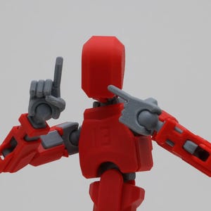 May include: A red and gray articulated robot toy with posable arms and legs. The robot has a red head and a gray body. The robot is in a pose with its arms raised and its legs spread apart.